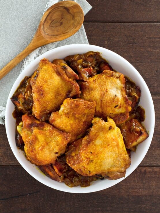 Bowl of Israeli Chicken Sofrito with potatoes, wooden spoon and cloth napkin on the side, on a wooden table.