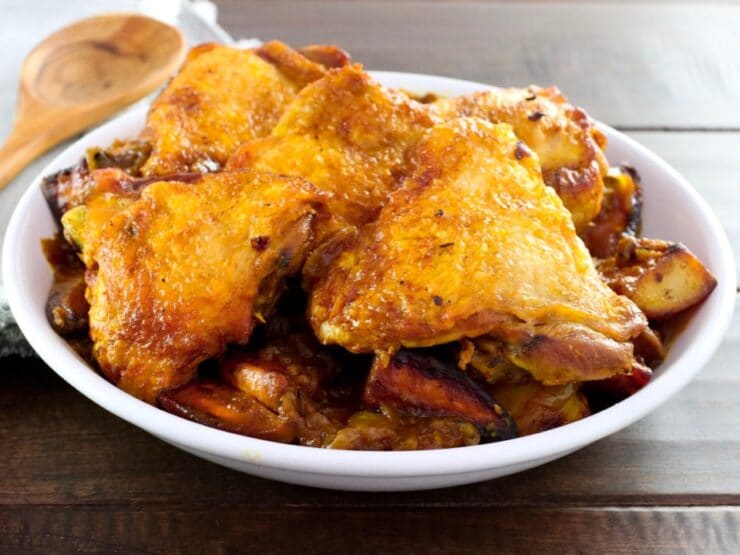 Israeli Chicken Sofrito - braised chicken pieces in a large shallow white bowl on wooden table with wooden spoon and napkin in background.