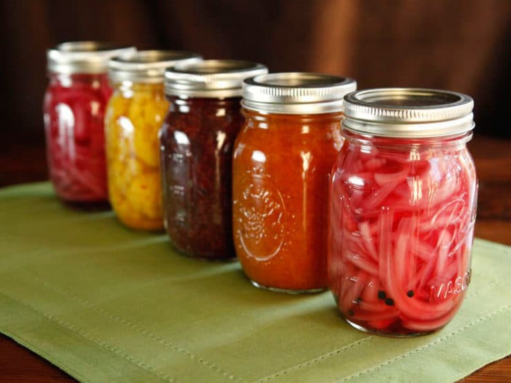 Five jars in a diagonal line, filled with canned colorful foods on a green placemat.