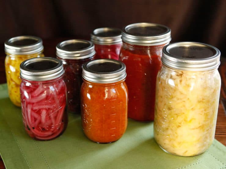 Seven jars staggered, filled with canned colorful foods on a green placemat.