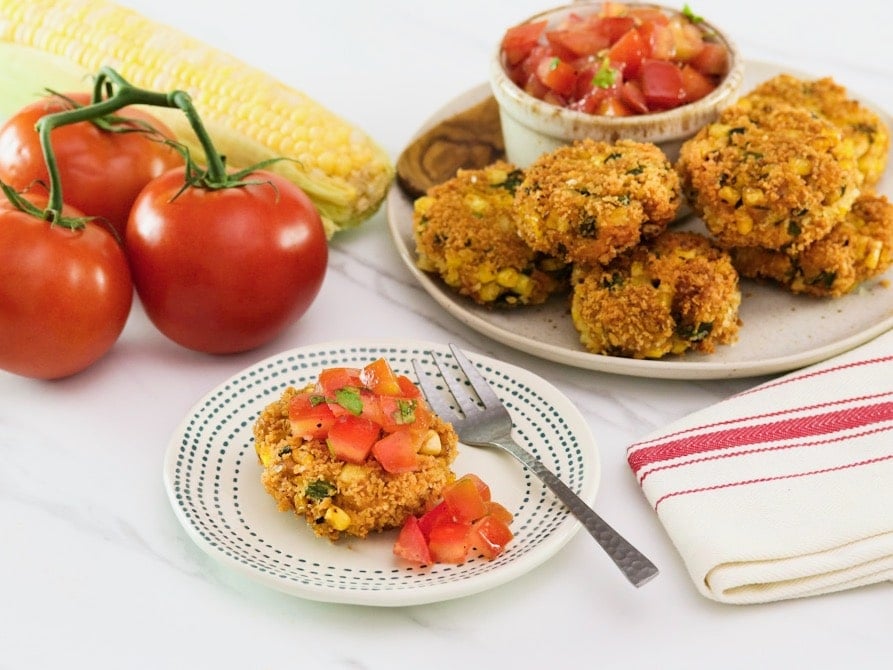 Horizontal shot of panko corn and pepper schnitzel, topped with tomato relish on a small plate with a fork. In the background there is a plate to the right with more schnitzel and relish, to the left there are whole tomatoes.
