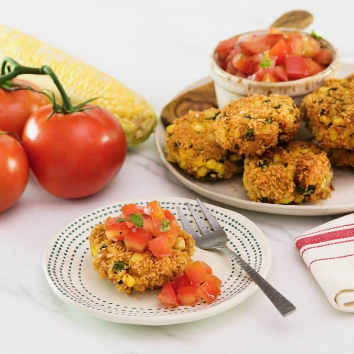 Panko corn and pepper schnitzel, topped with tomato relish on a small plate with a fork. In the background there is a plate to the right with more schnitzel and relish, to the left there are whole tomatoes.