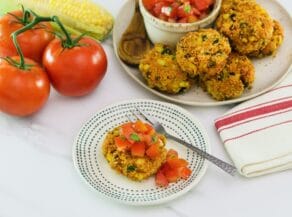 Horizontal angled overhead shot of panko corn and pepper schnitzel, topped with tomato relish on a small plate with a fork. In the background there is a plate to the right with more schnitzel and relish, to the left there are whole tomatoes.