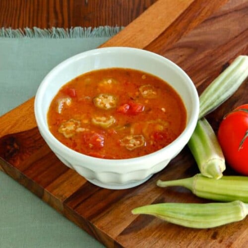 A horizontal shot of Okra soup served on a white bowl with fresh okra and tomato on side of the bowl