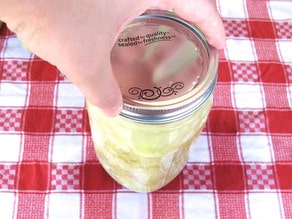 Hand placing lid onto jar to seal, red checkered towel in background.