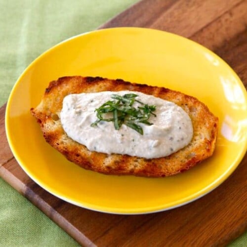 A toasted bread topped with Eggplant Tahini Basil Crostini served in a yellow plate on a wooden chopping board