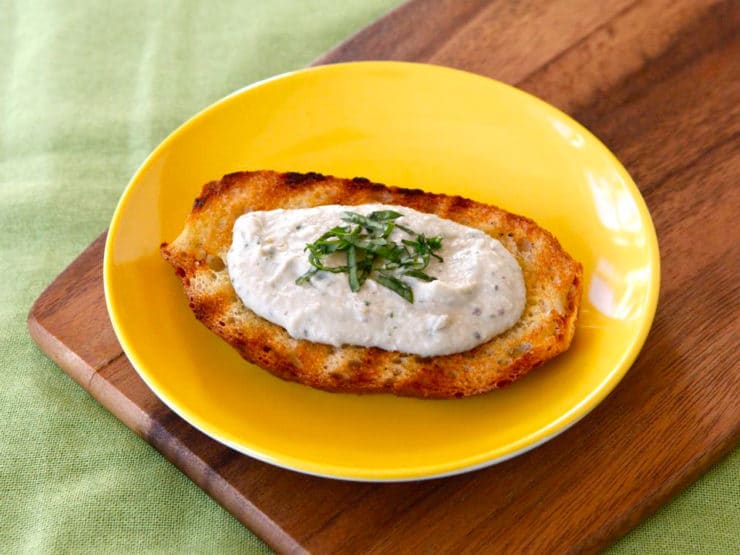 A toasted bread topped with Eggplant Tahini Basil Crostini served in a yellow plate on a wooden chopping board