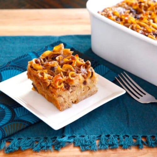 Rum Raisin Cranberry Kugel served on a small white square shaped plate with a casserole of Rum Raisin Cranberry Kugel and a fork on the side