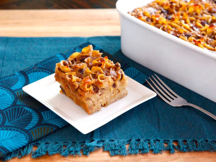 Rum Raisin Cranberry Kugel served on a small white square shaped plate with a casserole of Rum Raisin Cranberry Kugel and a fork on the side