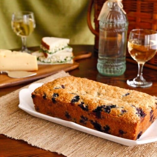 Blueberry Cake served on a white plate with a glass of win and a sliced cheese beside