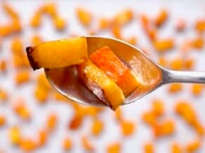 Close up of three pieces of caramelized roasted butternut squash in a metal spoon, with roasted butternut squash on a baking sheet soft focus in the background.