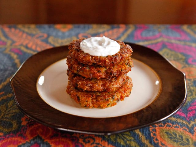 Pile of four curry vegetable latkes topped with sour cream on a white and brown plate atop a colorful placemat. 