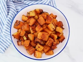 Overhead shot - large bowl of roasted butternut squash, caramelized. Dish sits on a white marble countertop with blue and white linen towel.