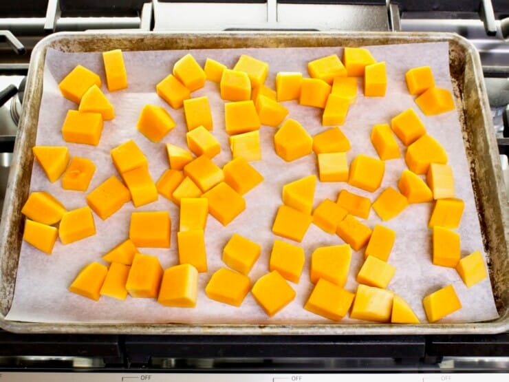 Overhead shot - tray of butternut squash cubes on a parchment-lined baking sheet, resting on a stovetop.