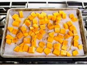 Overhead shot - tray of butternut squash cubes sprinkled with seasonings on a parchment-lined baking sheet, resting on a stovetop.