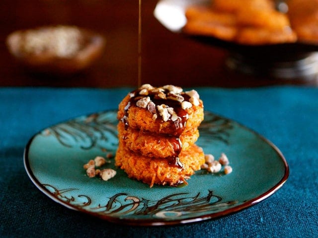 Pile of three orange sweet potato latkes drizzled with brown sugar syrup topped with candied nuts on green plate atop a teal placemat. 