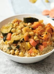 Square crop - dish of vegetable couscous on white marble background with towel, dry ingredients in backround - garlic, apricots, raisins.