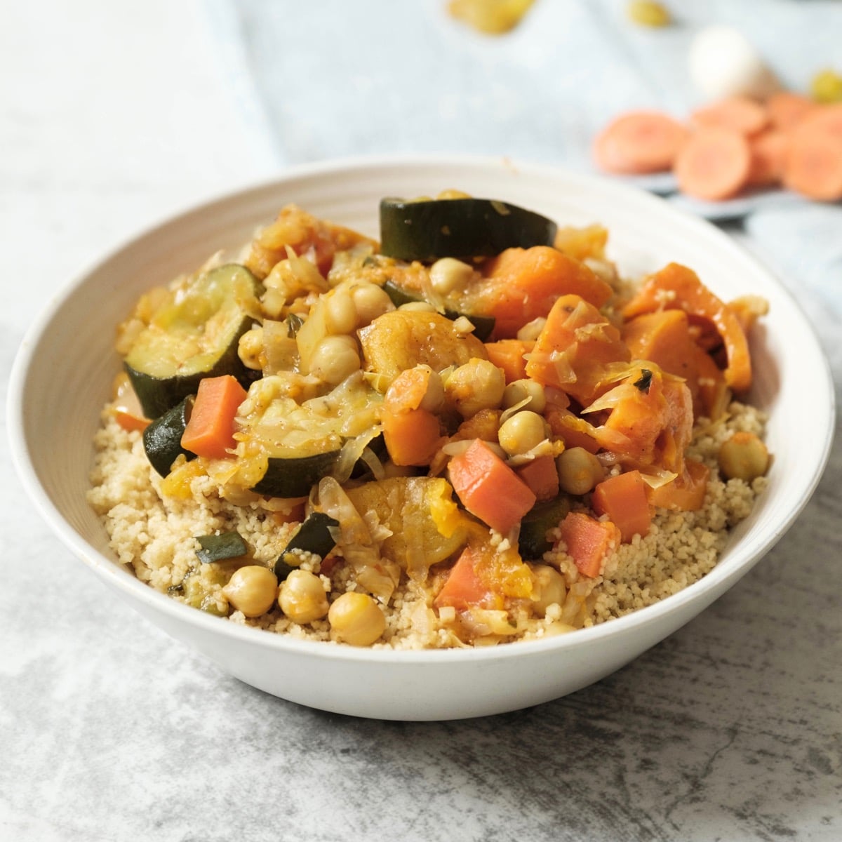 Square crop - dish of vegetable couscous on white marble background with towel, dry ingredients in backround - garlic, apricots, raisins.