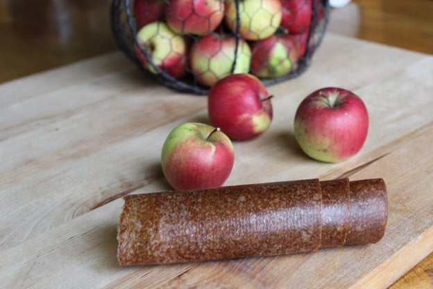 Rolled apple leather and three apples on a wooden chopping board and a set of apples on a container in the background