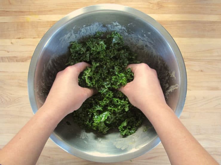 Two hands massaging kale with dressing in stainless steel bowl on wooden background.