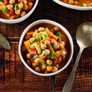 Overhead shot of chickpea chili garnished with cilantro with spoon on wooden table, two other bowls of chili on periphery.