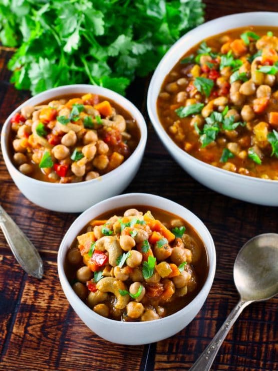 Vertical shot of two bowls of vegan chickpea chili with spoon and larger serving bowl of chili on wooden background, fresh cilantro in background.