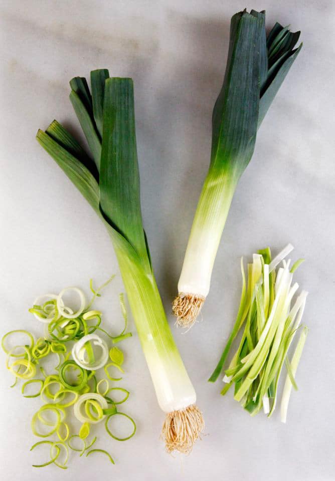 Two leeks on a marble board with julienne slices and rings of leek beside them. 