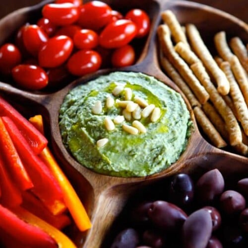 Close-up of white bean basil pesto hummus on wooden plate, surrounded by pine nuts, bell pepper, grape tomatoes, and breadsticks.