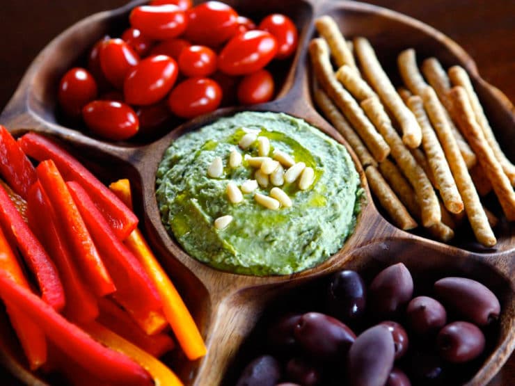 Close-up of white bean basil pesto hummus on wooden plate, surrounded by pine nuts, bell pepper, grape tomatoes, and breadsticks.