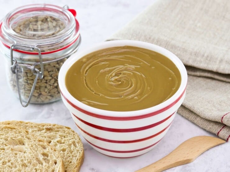 Horizontal shot - bowl of sunflower butter on a marble countertop with wooden spatula, bread slices, and cloth towel. Jar of sunflower seeds in background.