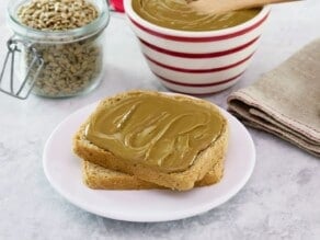 Two pieces of bread on a plate smeared with fresh homemade sunflower butter. Bowl of sun butter and jar of sunflower seeds in background, cloth napkin to the side.