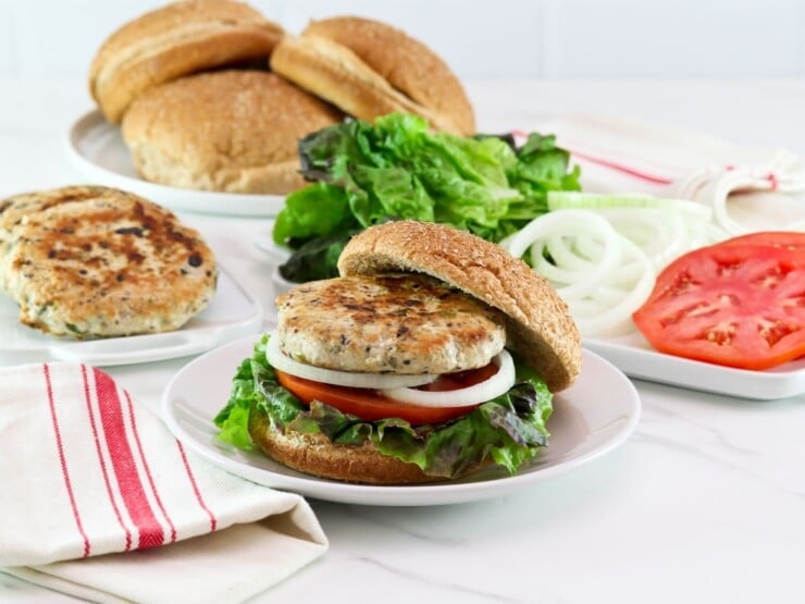Horizontal shot of a turkey burger on a bun with lettuce, tomato and onion. In the background there is a plate of buns, a plate of sliced tomato and onion, and a plate of turkey burger patties.