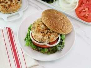 Horizontal overhead shot of a turkey burger on a bun with lettuce, tomato and onion. In the background there is a plate of buns, a plate of sliced tomato and onion, and a plate of turkey burger patties.