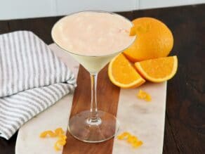 Close up of creamy orange creamsicle cocktail. Orange twist decorates the creamy froth. Citrus twists, orange with slices, and towel in background.