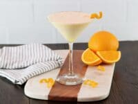 Horizontal shot of an orange creamsicle cocktail in a martini glass garnished with orange twist, on a wood and marble cutting board. Towel and orange with slices in background.