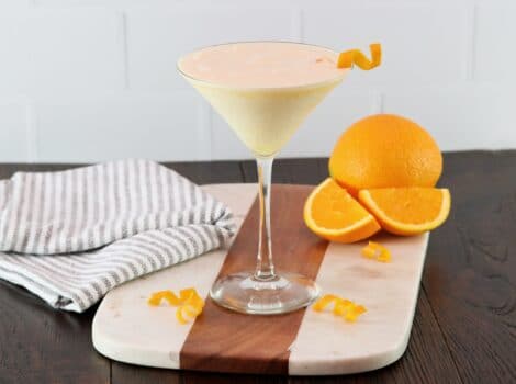 Horizontal shot of an orange creamsicle cocktail in a martini glass garnished with orange twist, on a wood and marble cutting board. Towel and orange with slices in background.