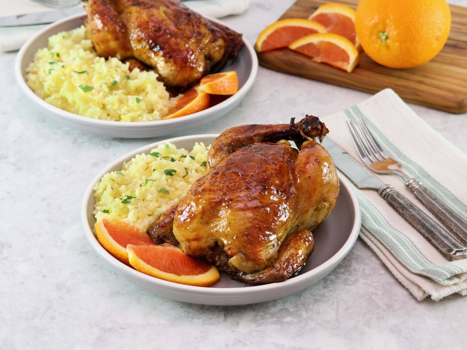 Horizontal shot of a plate containing a citrus marinated game hen on a bed of rice, garnished with fresh orange slices. Another plate of the same food sits in the background next to orange slices on a wooden cutting board.