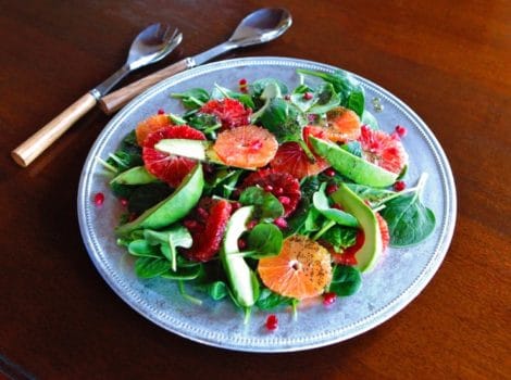 A vibrant citrus avocado salad with oranges, spinach, and pomegranate, drizzled with poppy seed dressing on a blue plate.