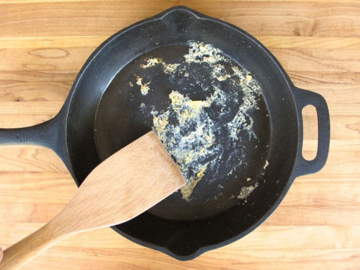 Scraping food from bottom of cast iron skillet with wooden spatula.