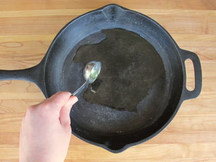 Hand drizzling oil into cast iron skillet with measuring spoon.