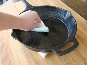 Hand wiping oil onto surface of cast iron skillet with paper towel.