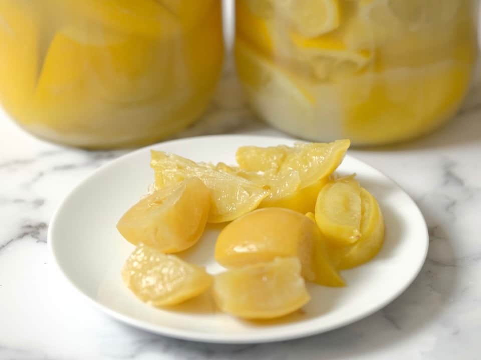 Dish of Preserved Lemon Quarters on marble counter, two jars of lemons in background.