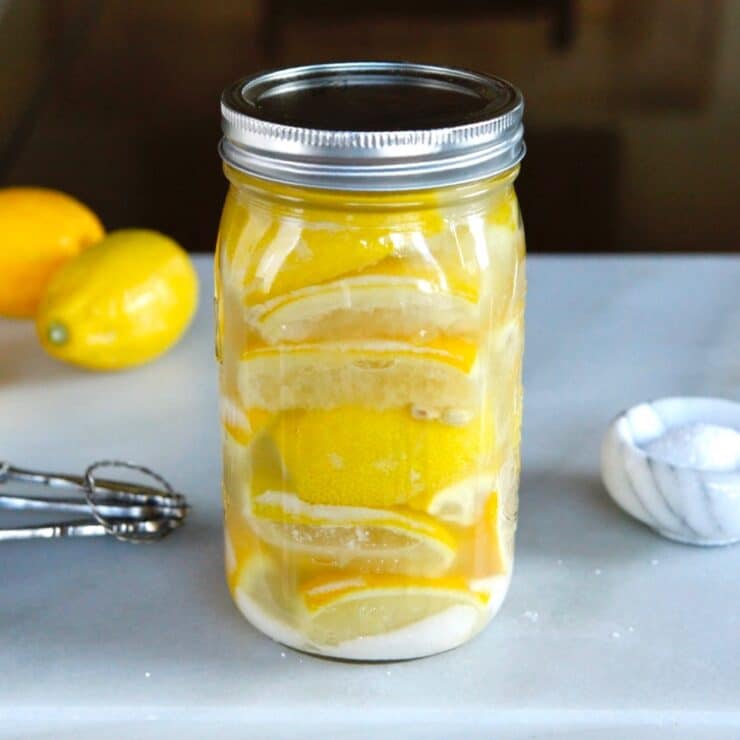 Jar of salt-preserved lemons on a marble cutting board with dish of salt, measuring spoons and lemons.