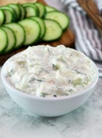 Horizontal shot - white bowl of cucumber raita with cucumber slices in the background.