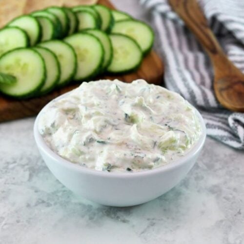Horizontal shot - white bowl of cucumber raita with cucumber slices in the background.