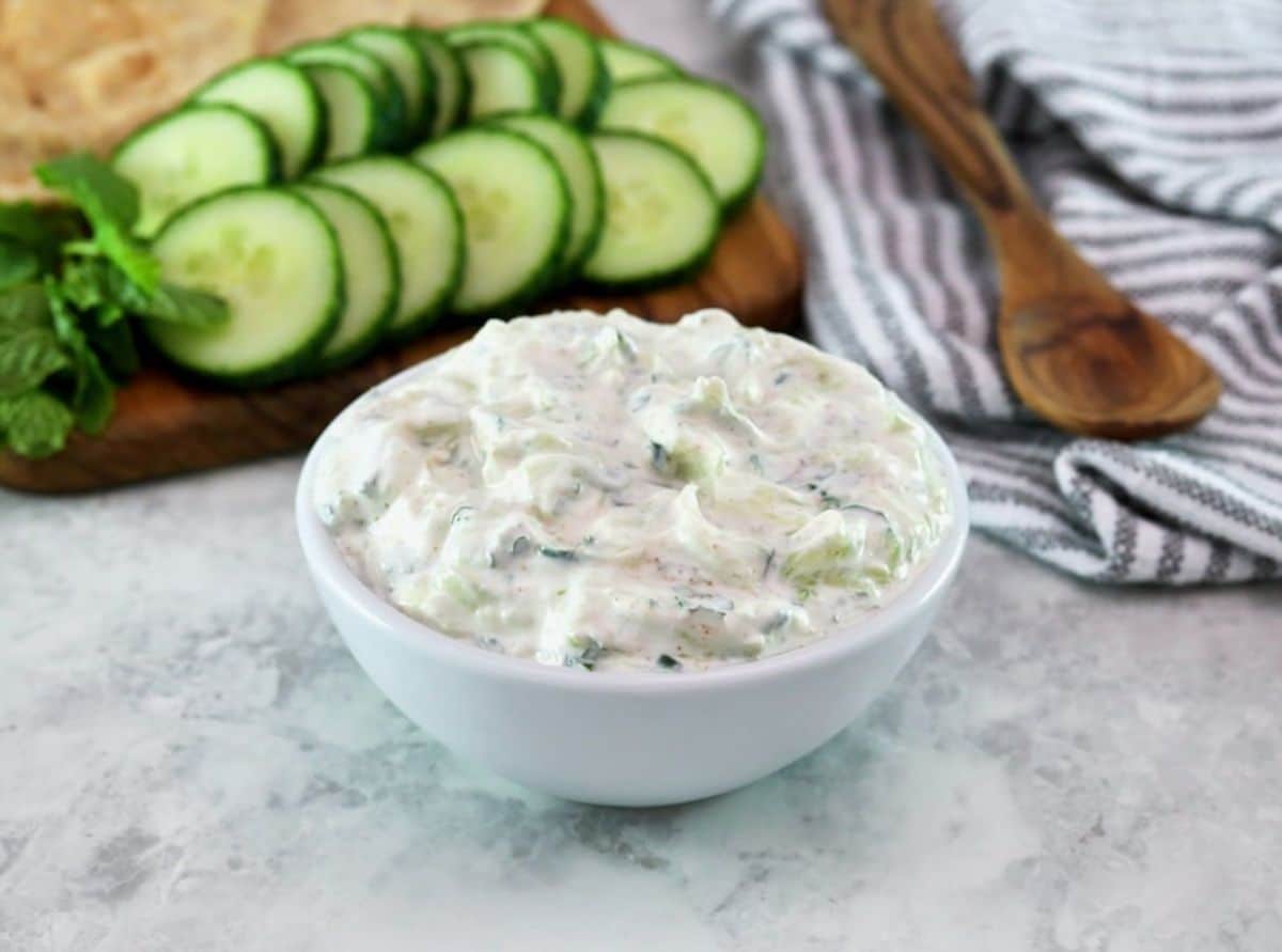 Horizontal shot - white bowl of cucumber raita with cucumber slices in the background.