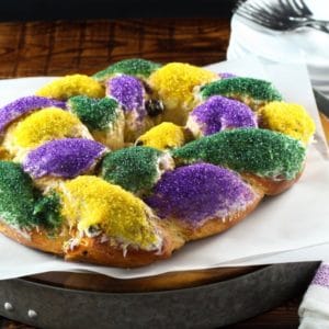 Brightly decorated braided circular King Cake on white parchment with towel in background.