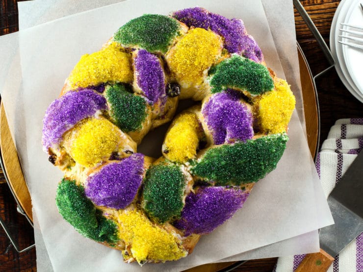 Overhead view of circular braided King Cake decorated with purple, yellow and green sprinkles on a white parchment paper sheet.
