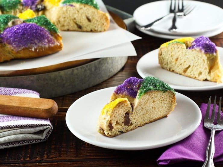 Colorful slice of King Cake on white plate with cake, plate and utensils in background.
