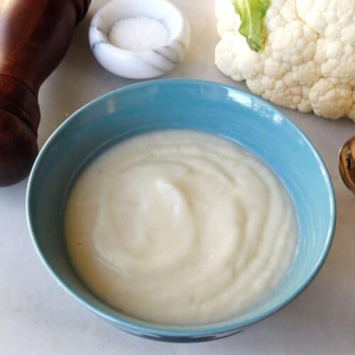 Image of a Cauliflower Soup on a bowl with a spoon, pepper crusher, salt, and cauliflower in the side